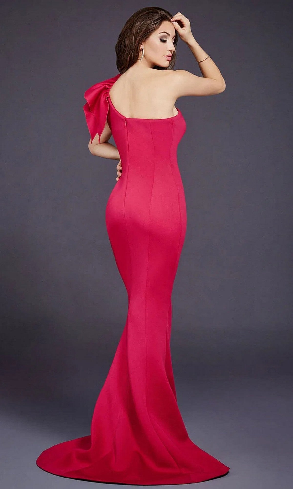 A woman with her back to the camera models the Jovani 32602, a one-shoulder mermaid prom gown in pink with a shoulder bow, against a plain gray background.